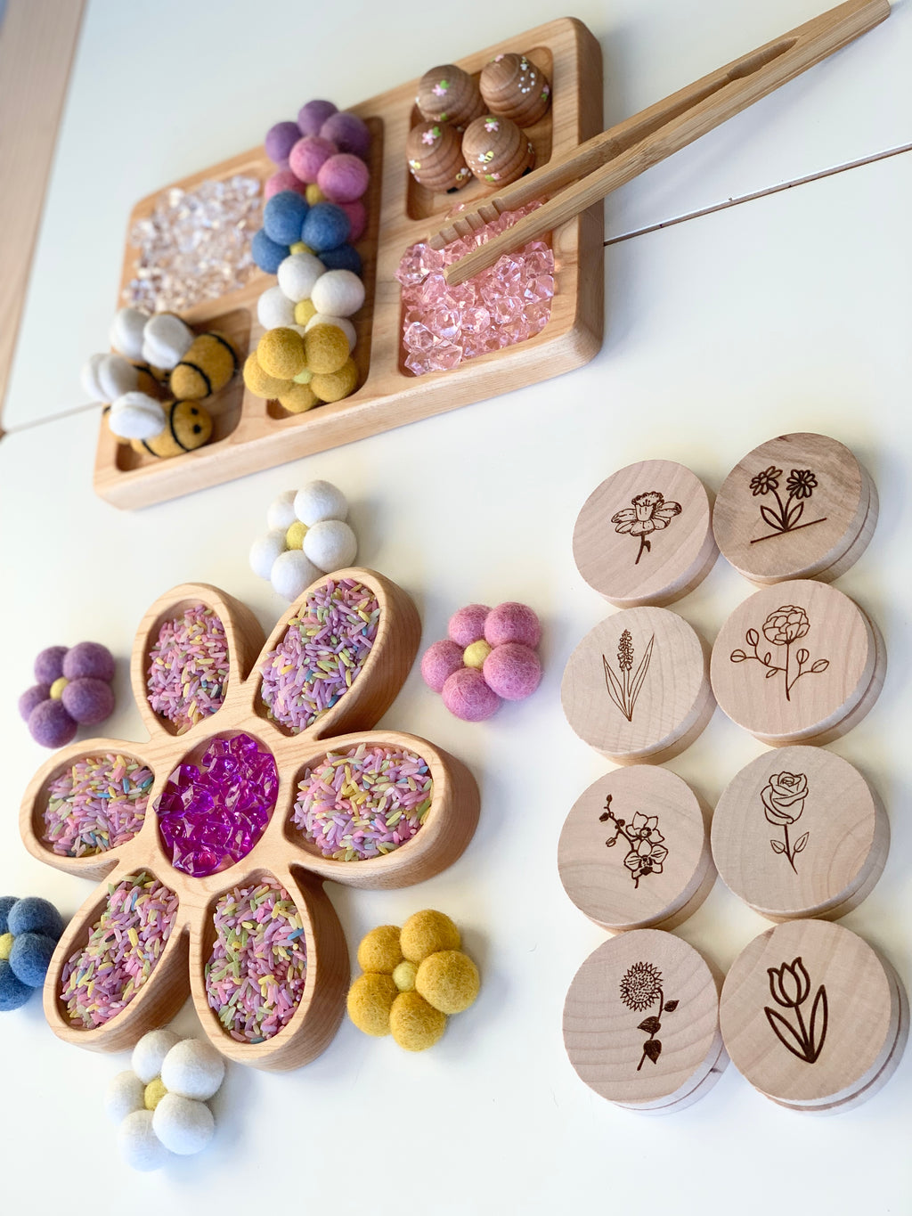 Natural wooden rectangle tray and flower-shaped tinker tray filled with colorful felt balls, rice, and pink gems on white surface