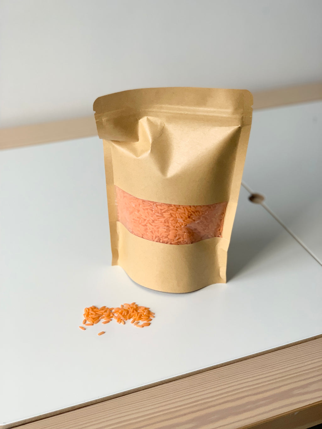 Brown kraft resealable pouch filled with orange sensory rice with some spilled grains on white surface