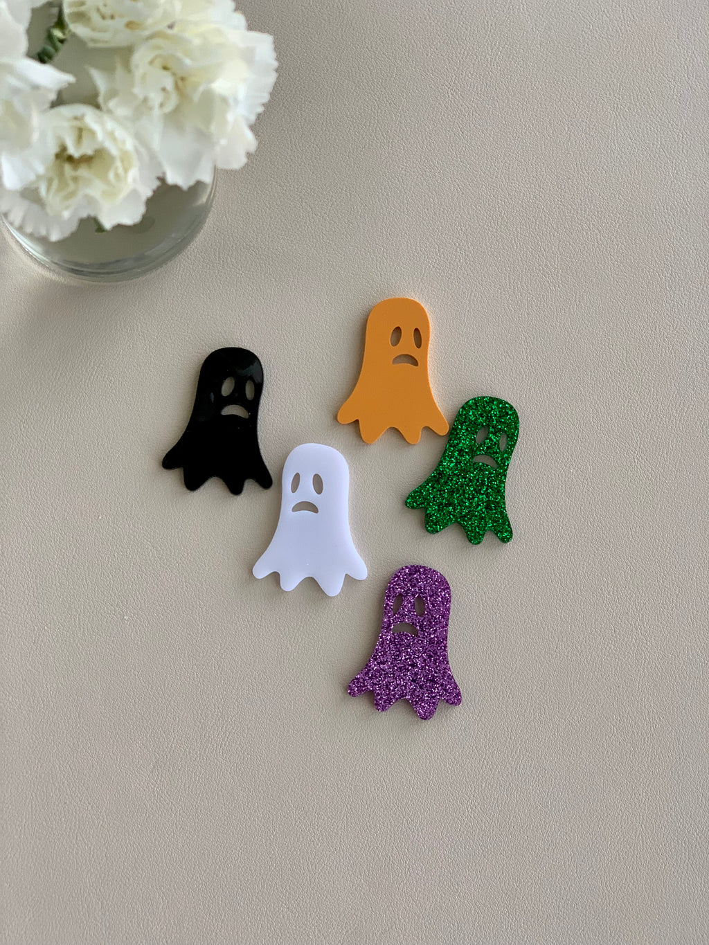 Set of five small laser-cut acrylic ghost shapes in black, orange, white, green glitter, and purple glitter on neutral surface