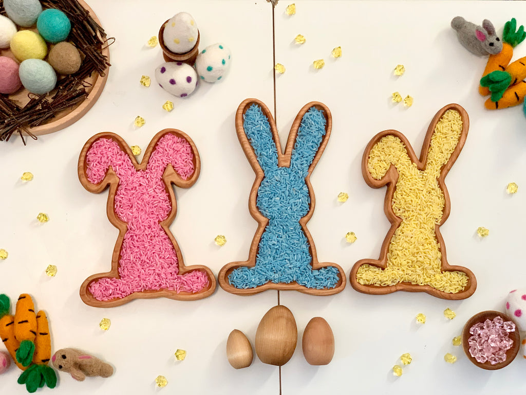Three wooden bunny-shaped sensory trays filled with pink, blue, and yellow rice surrounded by Easter-themed decorations