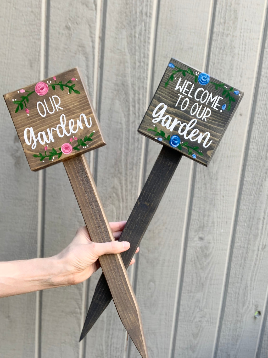 Two small square wooden garden signs with white lettering and floral accents on wood stakes held by a hand