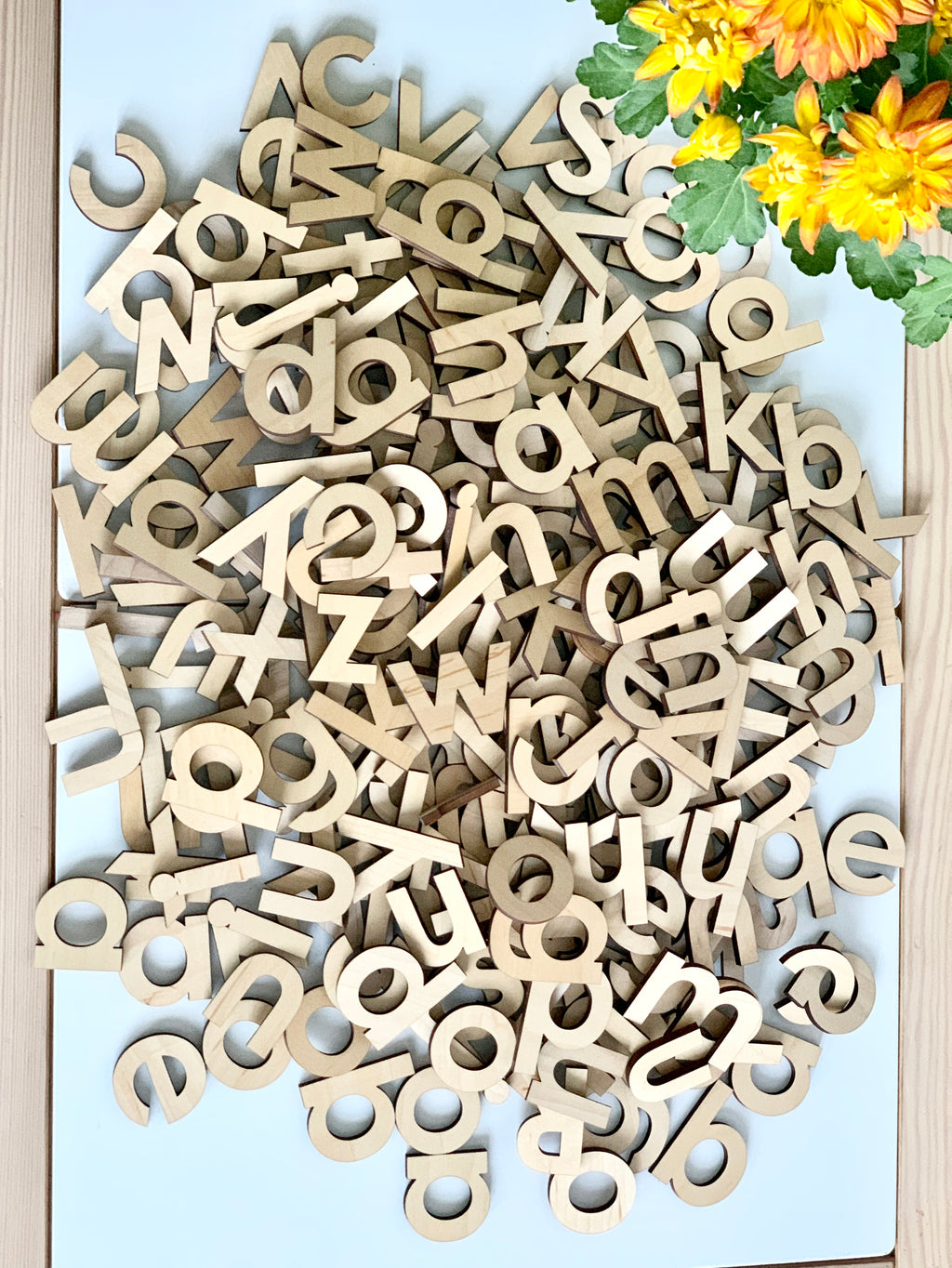 Natural wood lowercase alphabet letters scattered on white surface with orange and yellow flowers in corner