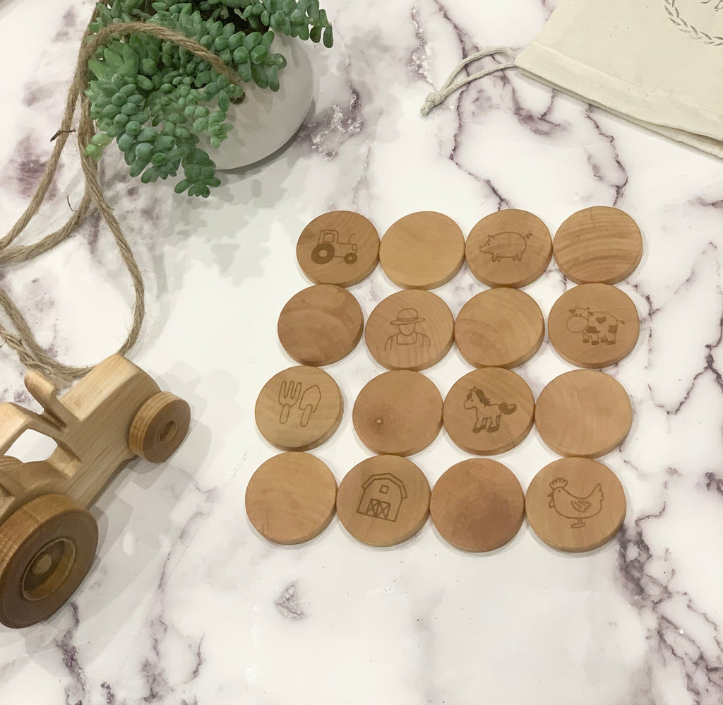 Natural wooden memory game pieces with engraved farm illustrations arranged on white marble surface next to wooden tractor toy