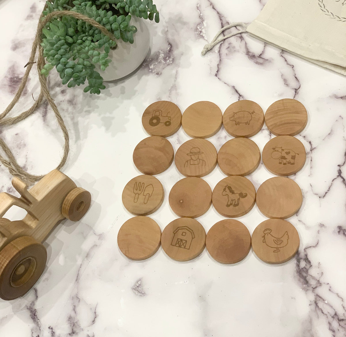 Natural wooden memory game pieces with engraved farm illustrations arranged on white marble surface next to wooden tractor toy