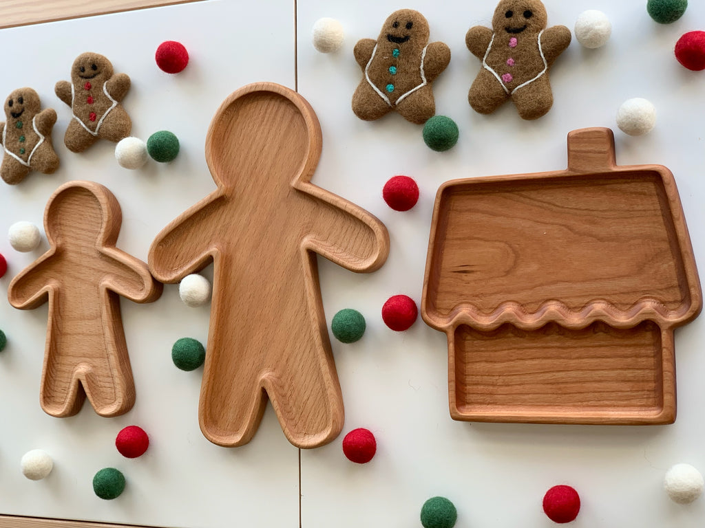 Three wooden sensory trays shaped like gingerbread people and a house surrounded by felt balls on white surface