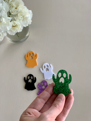Set of five laser cut acrylic ghost shapes in orange, white, black, purple glitter, and green glitter on beige surface