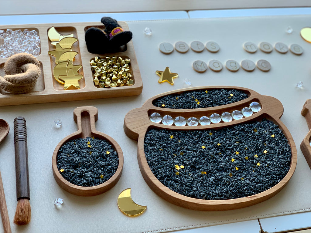 Wooden sensory play trays filled with black dyed rice and gold star confetti with wooden magic-themed accessories