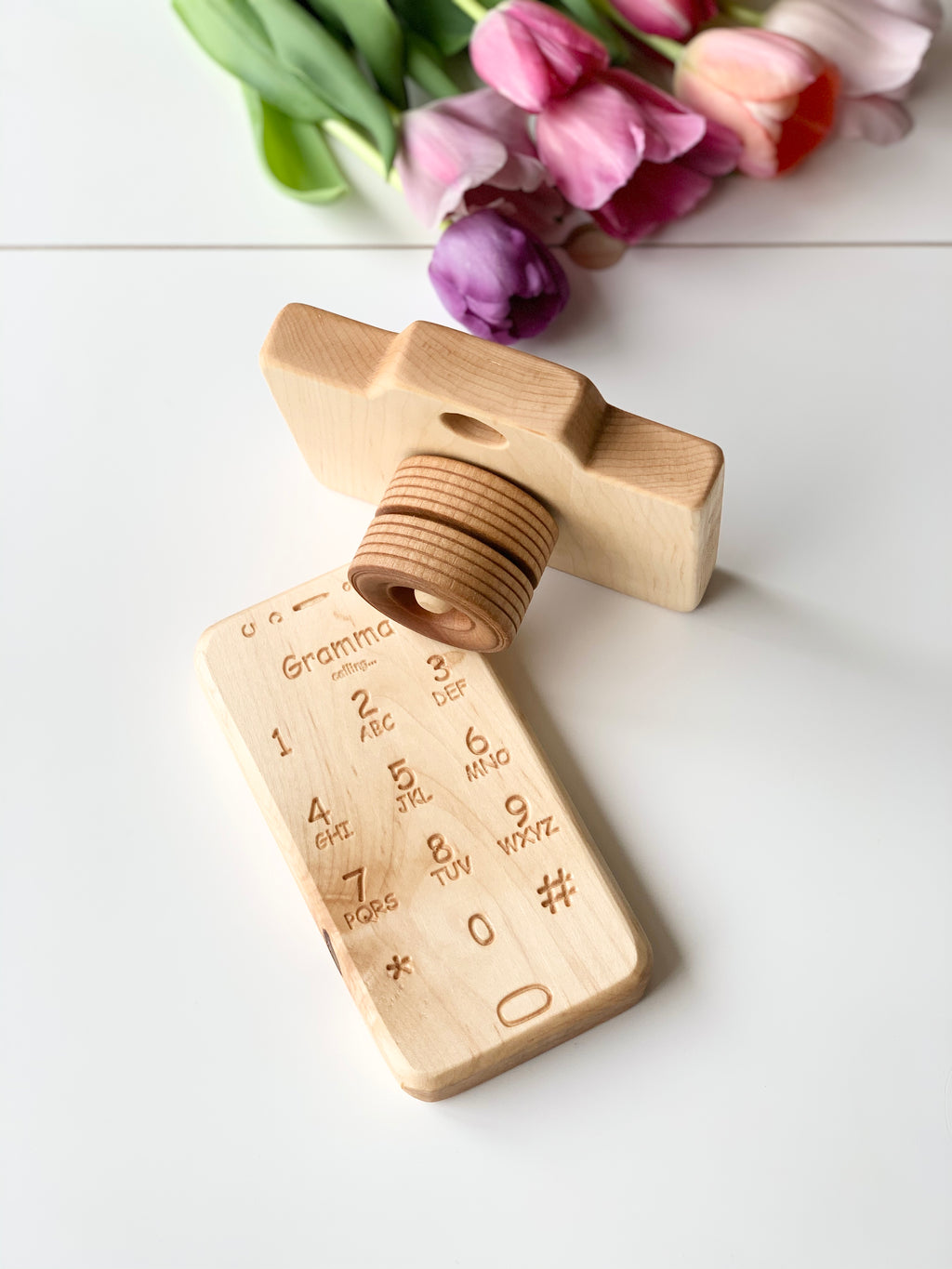 Natural wooden toy camera with engraved rotating lens and wooden toy phone with engraved numbers on white surface with tulips