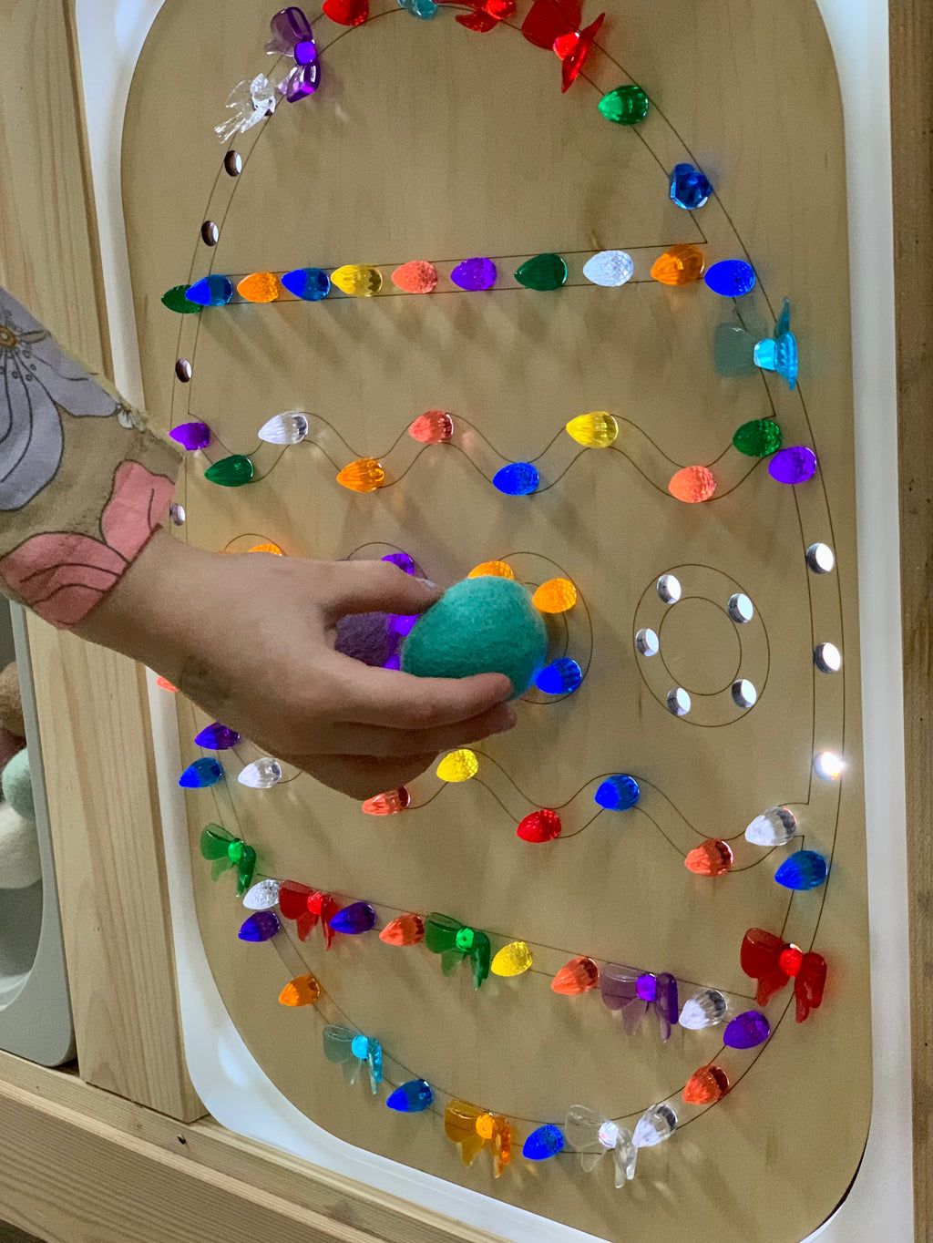 Child's hand placing green felt egg on wooden geometric board with colorful light-up pegs in various shapes
