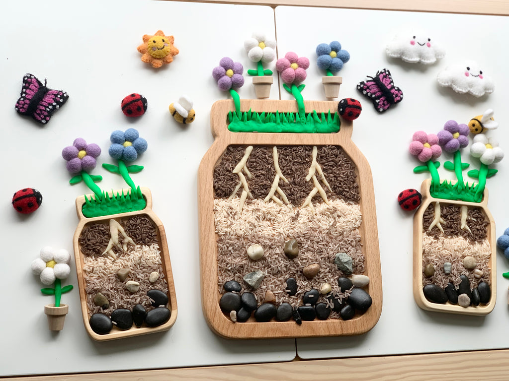 Three large jar-shaped sensory trays with layered natural materials and felt flower and insect decorations on white surface