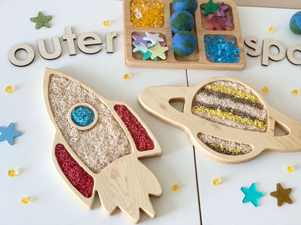 Natural wooden sensory tray set shaped like a rocket and Saturn planet filled with colored rice and grains on white surface