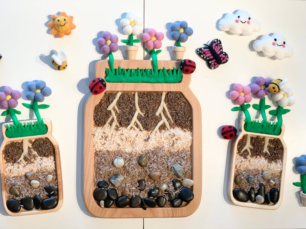 Large jar plate sensory tray with soil layers, roots, rocks, felt flowers, ladybugs, bees, butterfly, and smiling clouds on white background