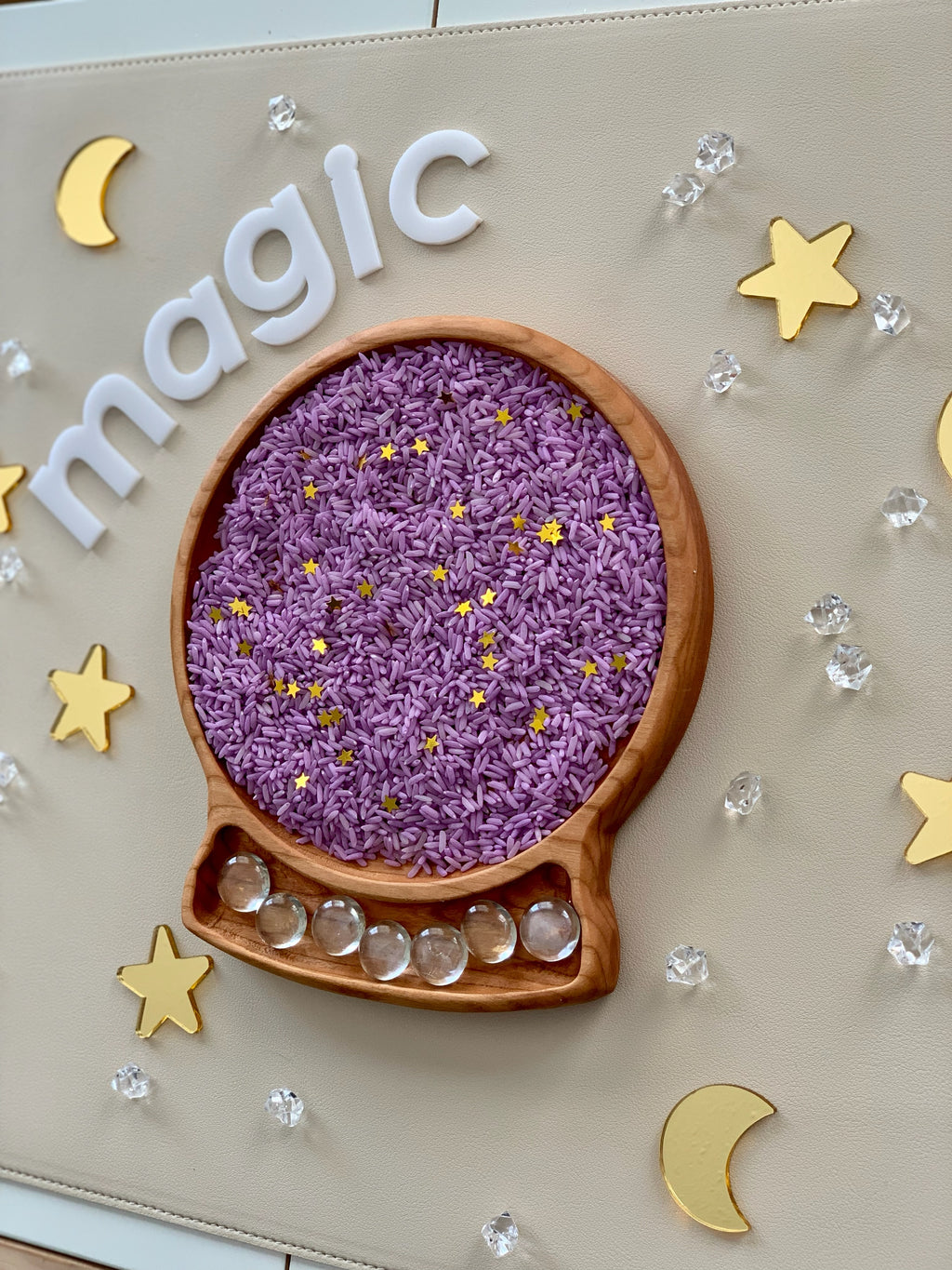 Wooden tray shaped like a crystal ball filled with purple rice and gold star acrylic shapes on beige surface with clear beads