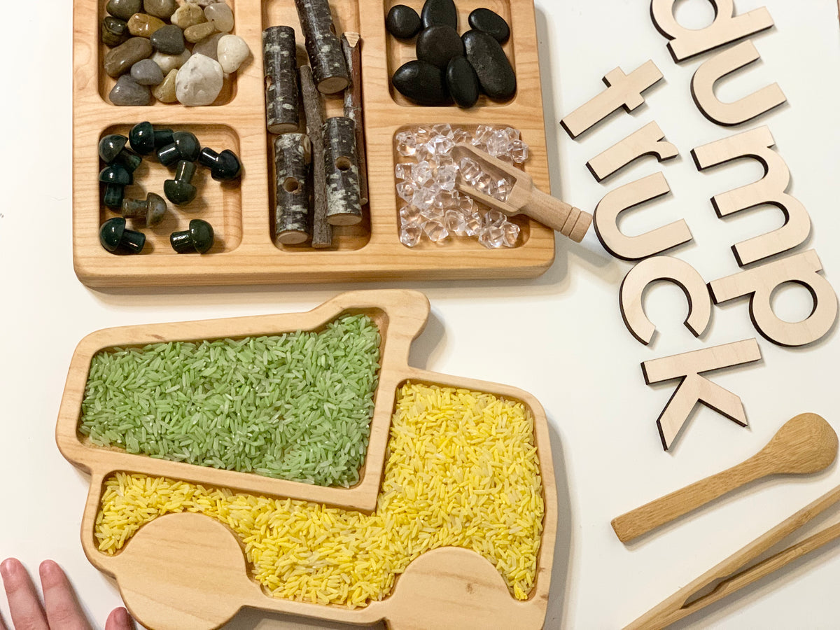 Wooden dump truck shaped sensory tray filled with colored rice, natural stones, and wooden scoop on white surface