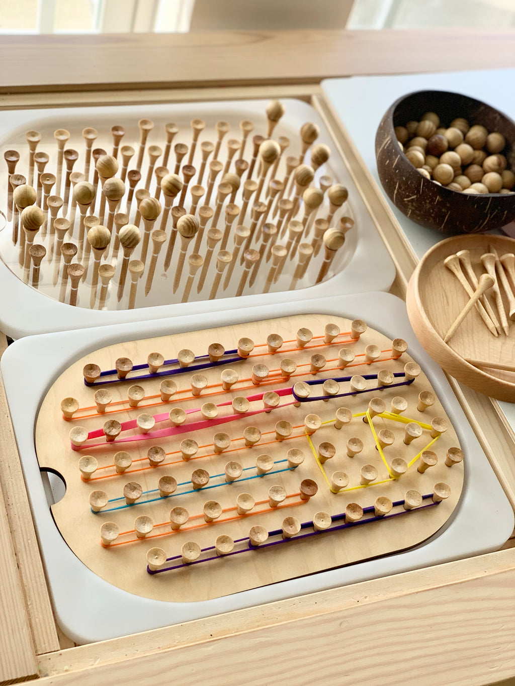 Wooden geometric peg board insert with small pegs and colorful elastic bands on light wood tabletop with bowls of pegs nearby