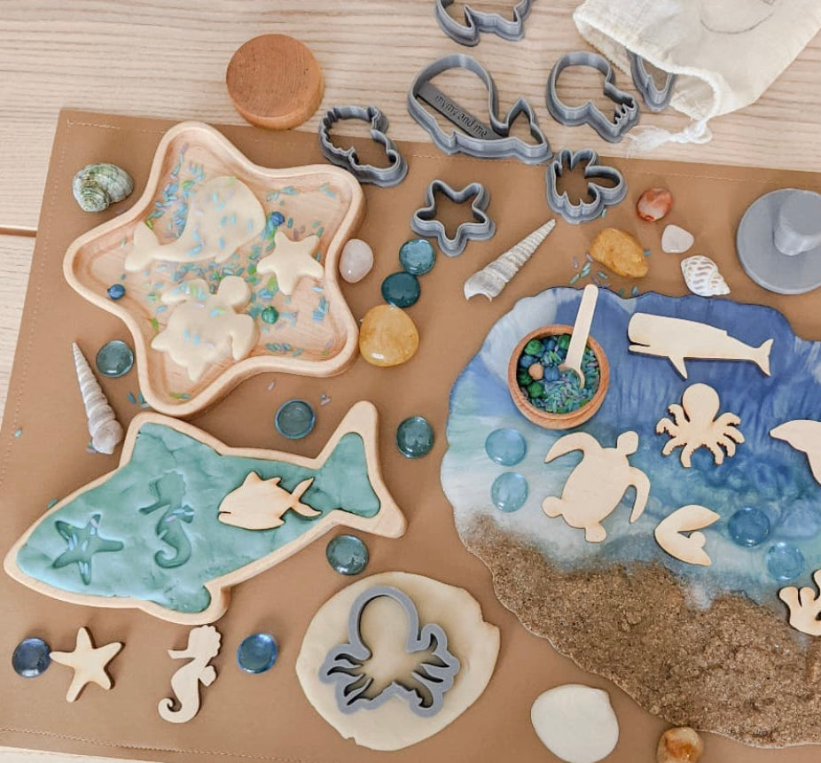 Wooden star and fish-shaped sensory trays with sea creature cutouts, shells, and glass pebbles on craft paper background