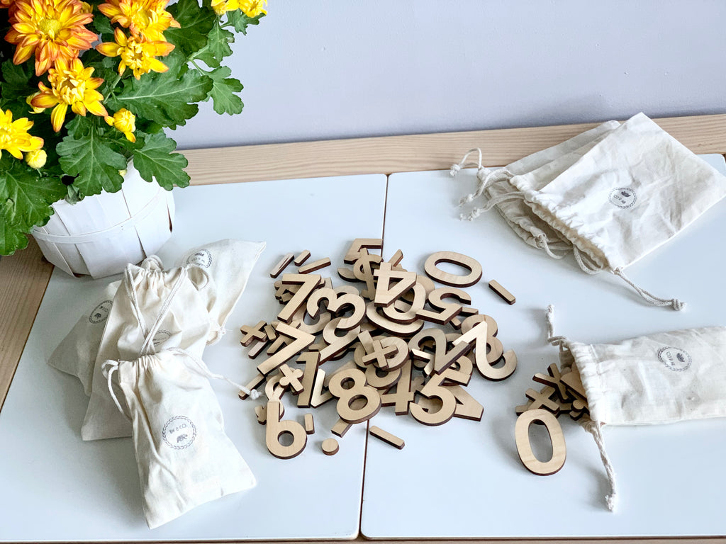 Maple wooden number set with numerals and math symbols scattered on white surface next to cloth storage bags and yellow flowers