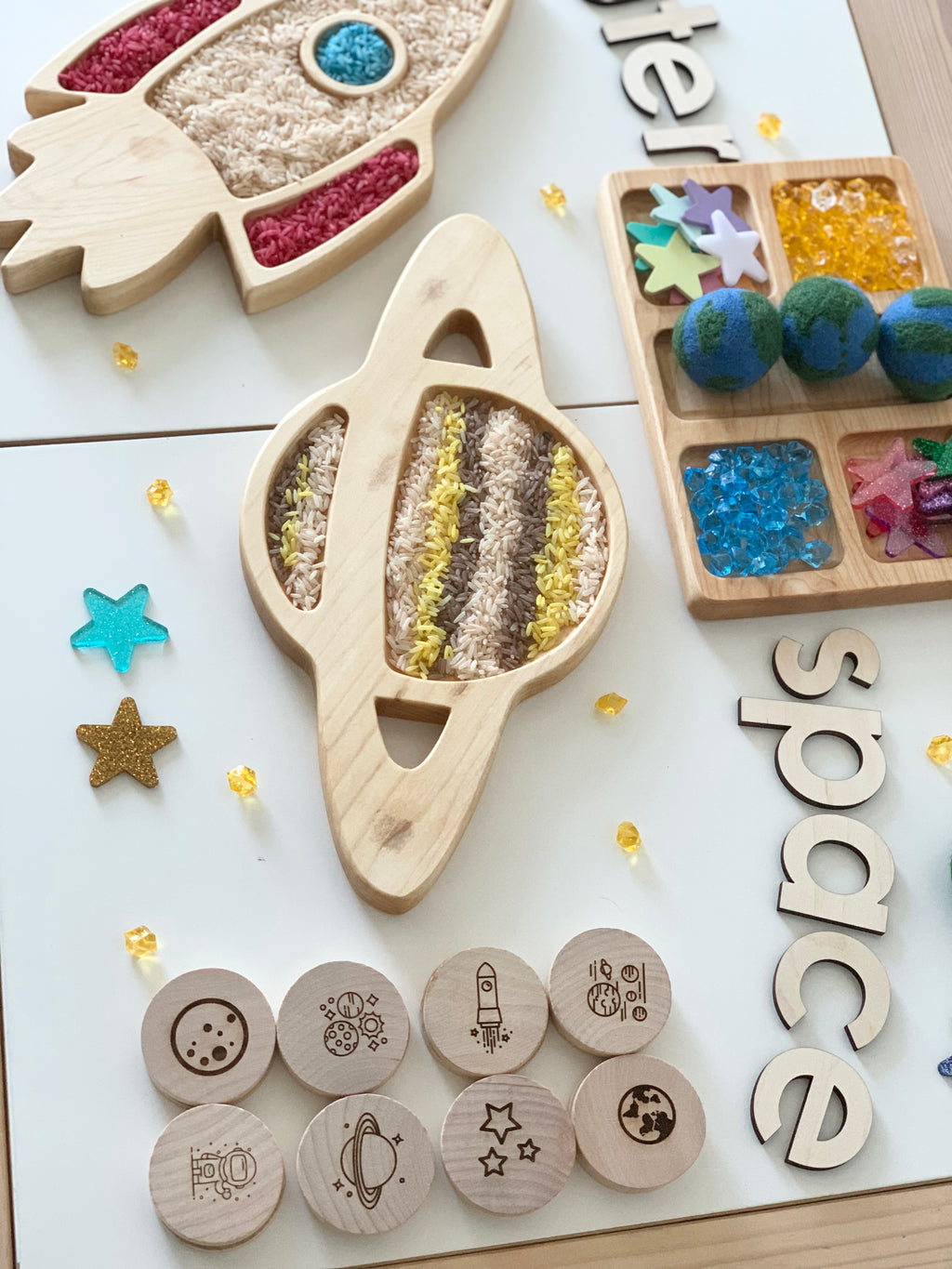 Natural wooden Saturn planet sensory tray with colored rice, surrounded by wooden space-themed tokens and decorative stars on white surface