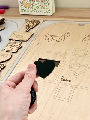 Hand holding black acrylic heart shape over engraved wooden sensory play board with love-themed outlines