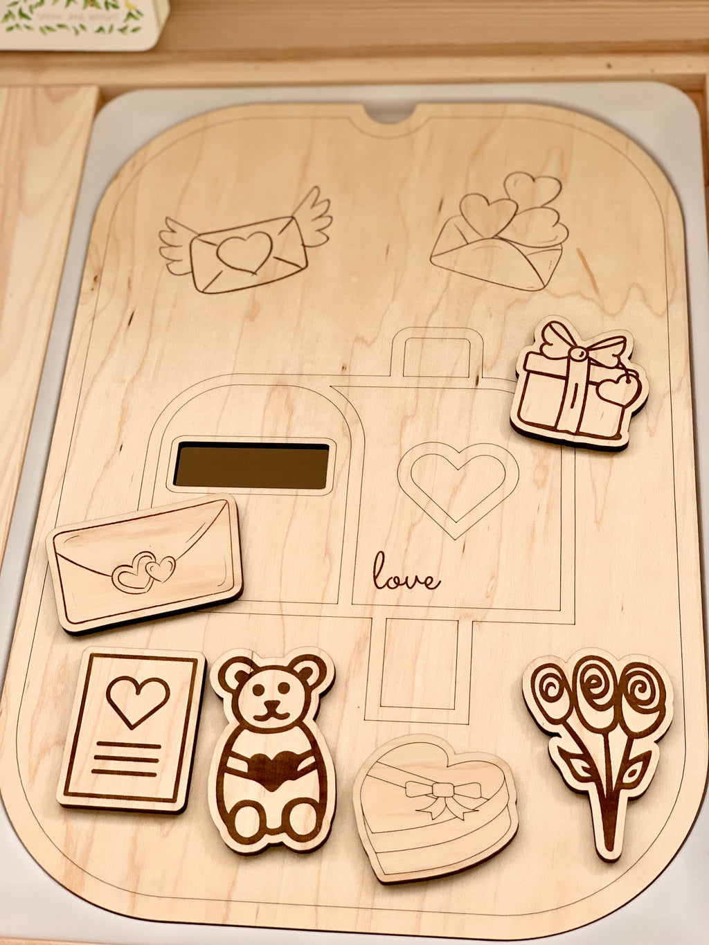 Wooden love post mailbox tabletop insert with engraved outlines and six small wooden Valentine-themed shapes on light wood surface