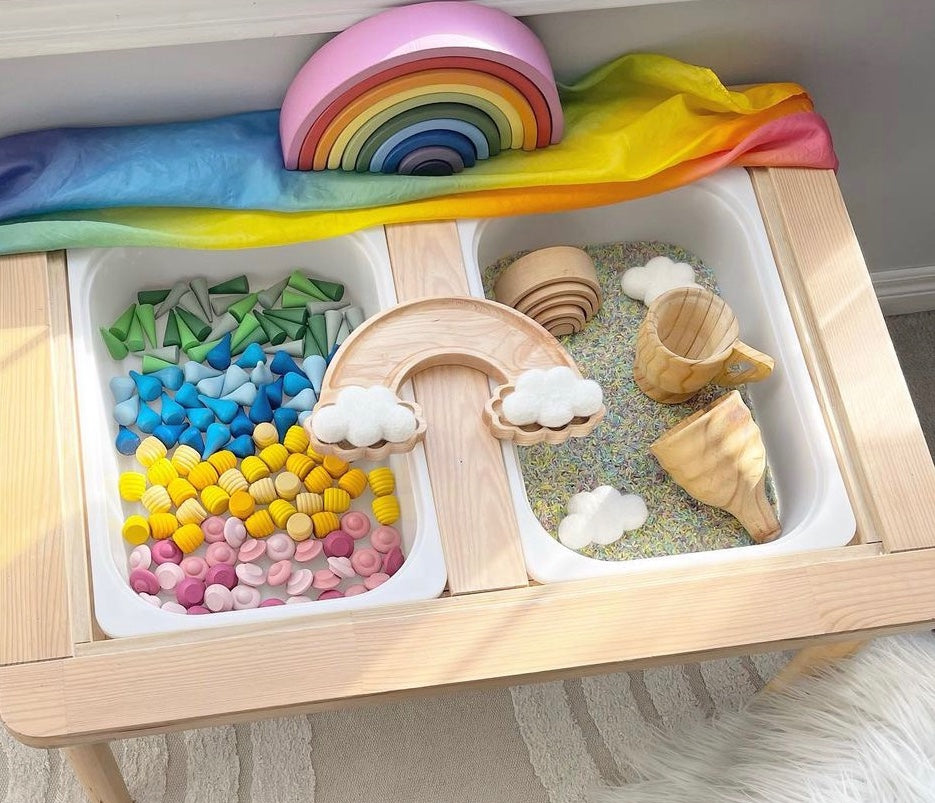 Wooden sensory play tray with colorful sensory beads, wooden rainbow arch, cup, and spiral toy on a light wood table
