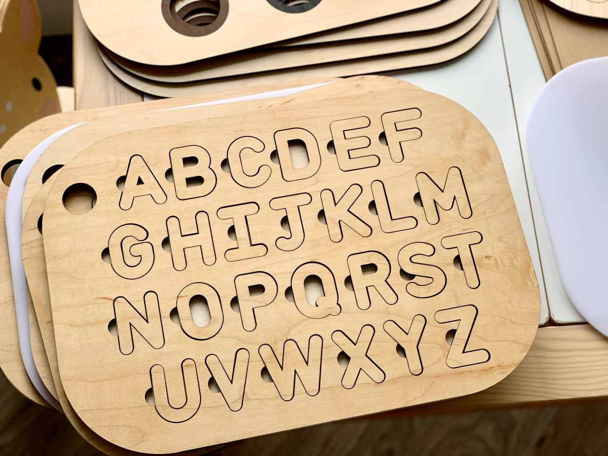 Maple plywood uppercase alphabet puzzle insert with cut-out letters on wooden tabletop, stacked with other inserts beneath