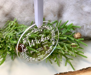 Round clear acrylic Christmas ornament engraved with family names and decorative leafy wreath hanging on white ribbon