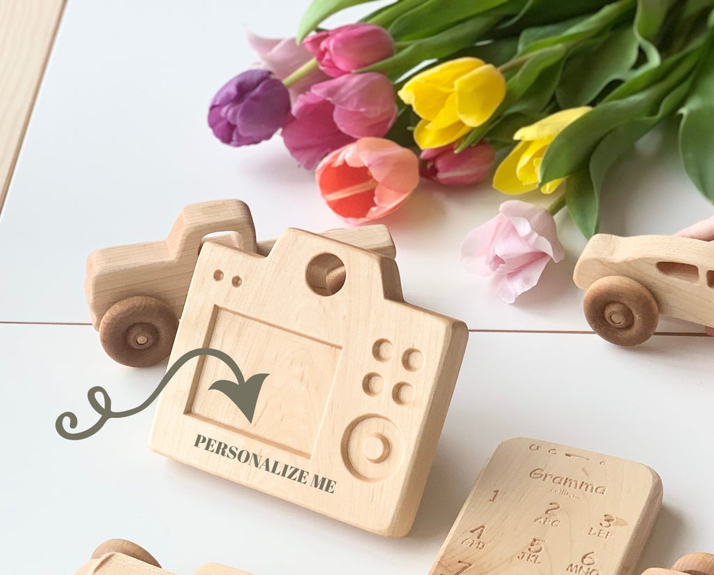 Natural wooden toy camera with engraved buttons and rotating lens surrounded by wooden cars and colorful tulips on table