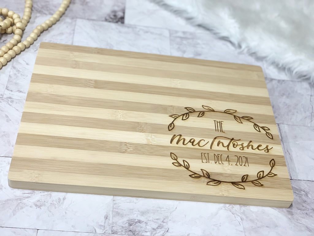 Rectangular striped bamboo cutting board with personalized engraved leaf wreath and family name in bottom right corner