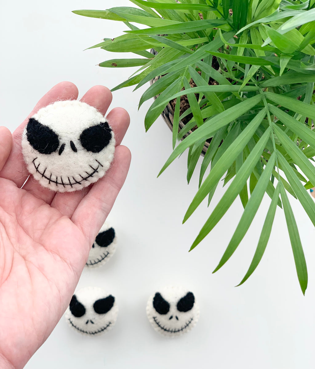 Hand holding one small handmade white felt Halloween skull with black stitched eyes and smile near green plant on white background
