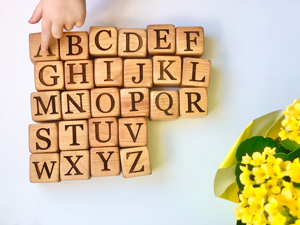Natural wooden 1.5-inch alphabet blocks with engraved capital letters arranged in rows on white surface with child's hand and yellow flowers