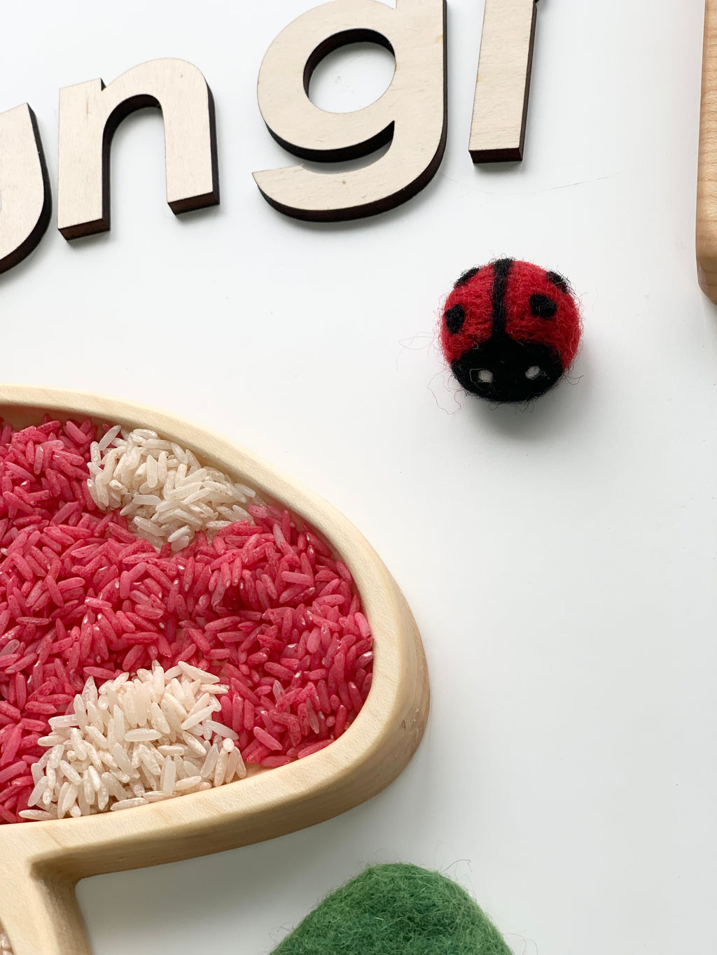 Small handmade red and black felt ladybug with white eyes on white background near wooden letters and colored rice in wood tray