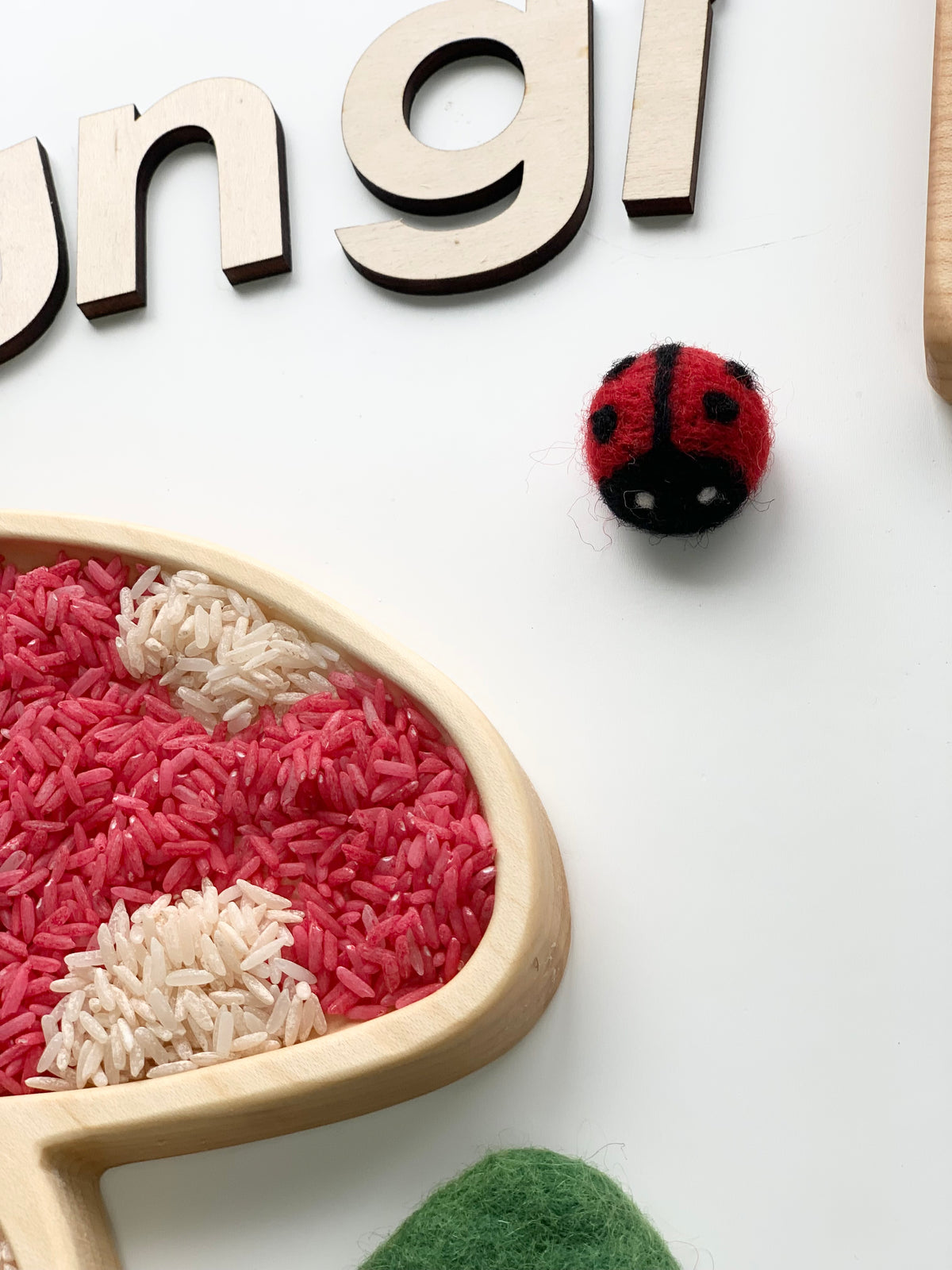 Small handmade red and black felt ladybug with white eyes on white background near wooden letters and colored rice in wood tray