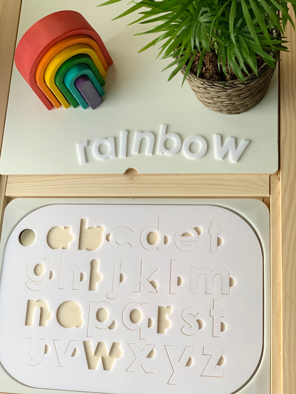Lowercase alphabet puzzle insert with white acrylic letters on maple plywood base next to colorful wooden rainbow and potted plant