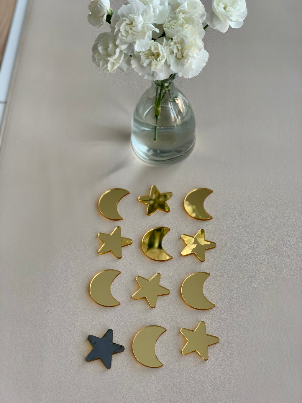 Set of twelve glossy gold acrylic moon and star shapes arranged in rows on beige surface with white flowers in glass vase
