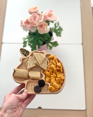 Natural wooden unicorn-shaped sensory tray with snacks including crackers, goldfish, cookies, and seeds held over white table with flowers