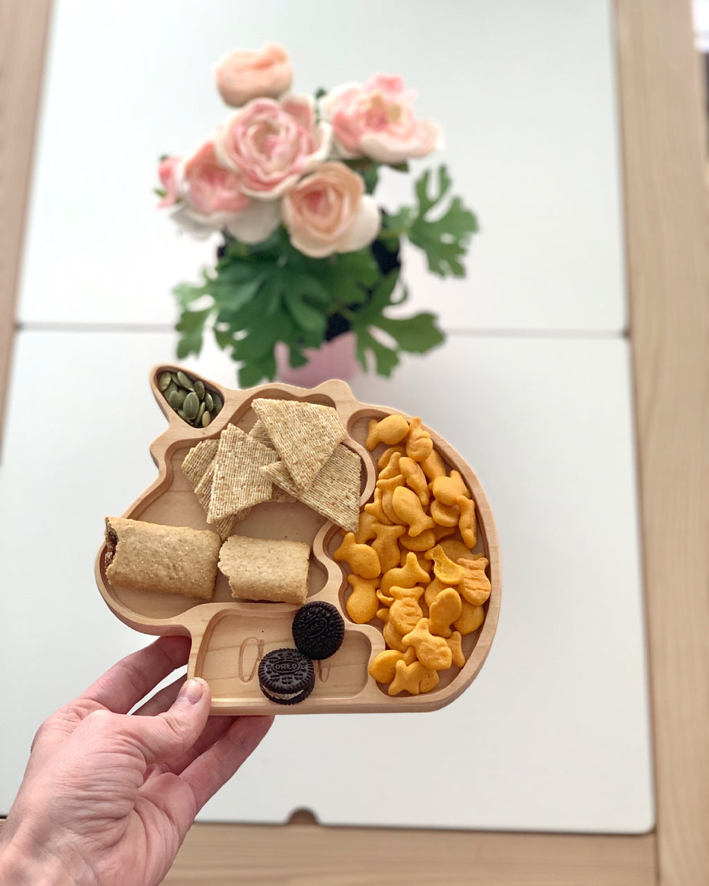 Natural wooden unicorn-shaped sensory tray with snacks including crackers, goldfish, cookies, and seeds held over white table with flowers