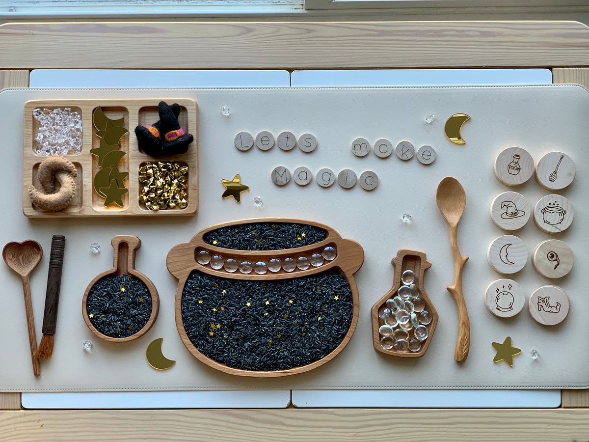 Wooden sensory tray set with cauldron plate filled with black rice, wooden letter beads spelling lets make magic, and themed wooden tokens