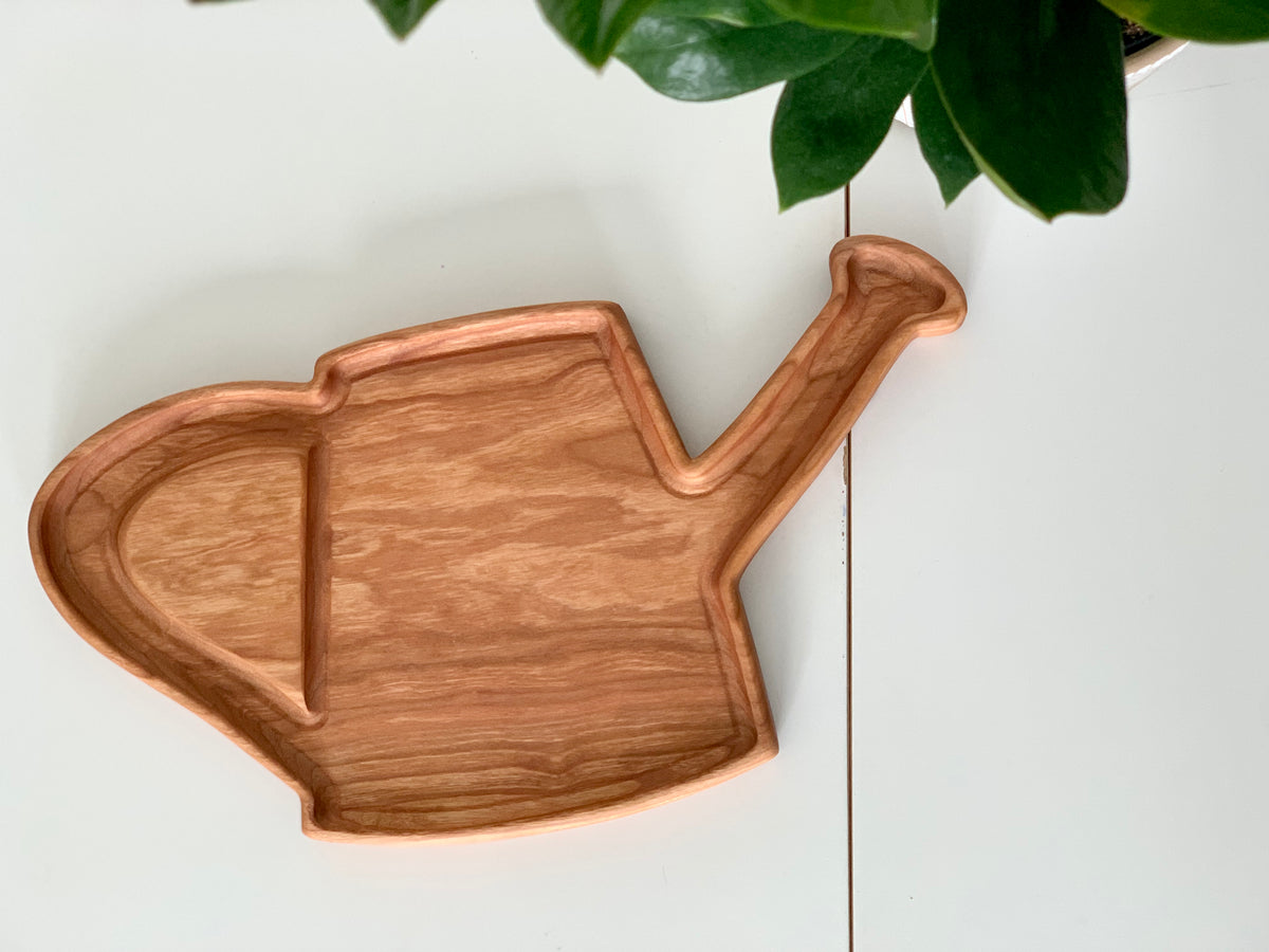 Wooden sensory tray in the shape of a watering can on white surface with green plant leaves nearby