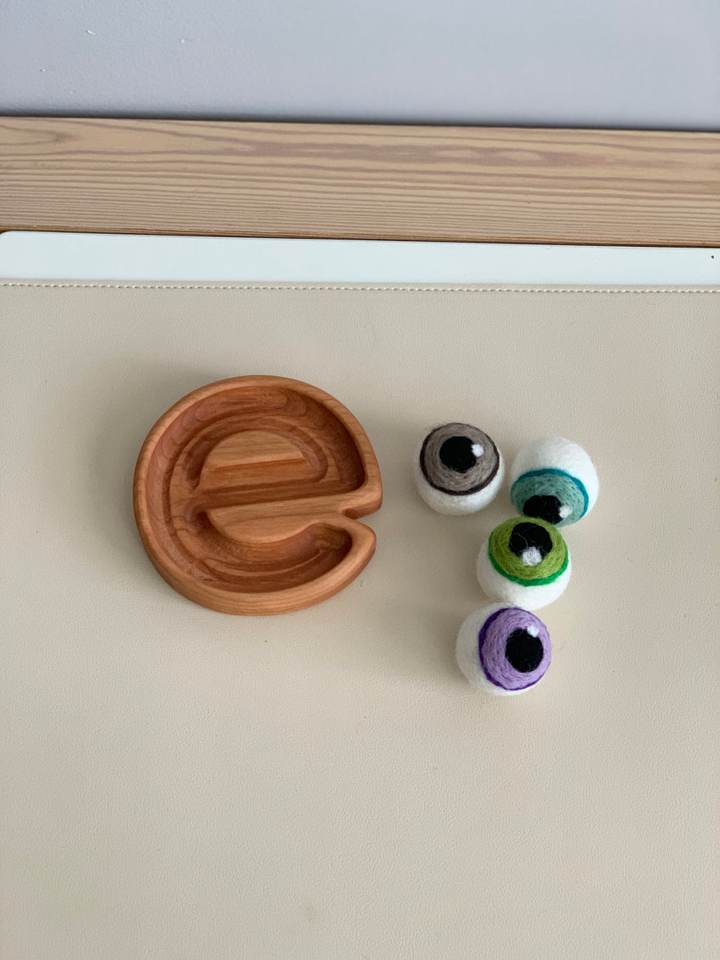Small wooden letter e sensory tray with four colorful felt balls on cream surface and wooden edge background