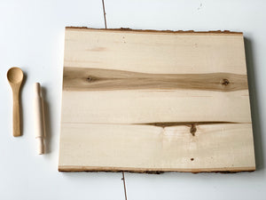 Live edge wooden tray with natural bark edges and wood grain, next to small wooden spoon and rolling pin on white surface