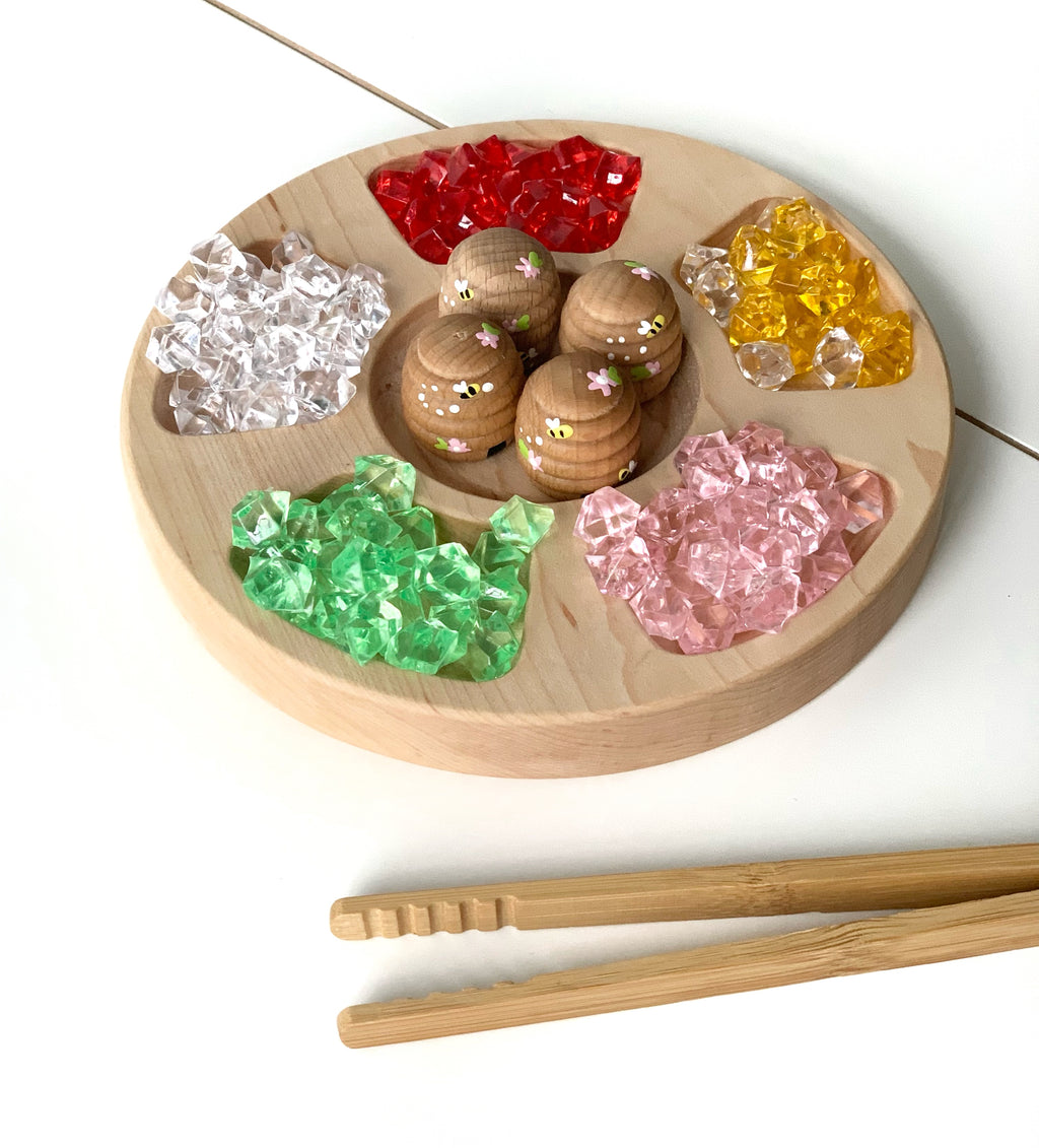 Round wooden tinker tray with five compartments filled with colorful translucent gems and wooden peg dolls on white background
