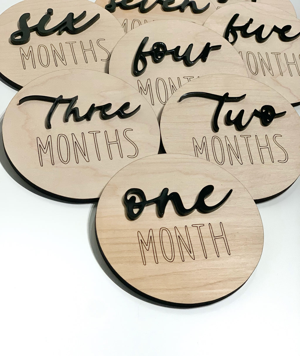 Round wooden monthly milestone discs with black cursive numbers and engraved text on light wood background