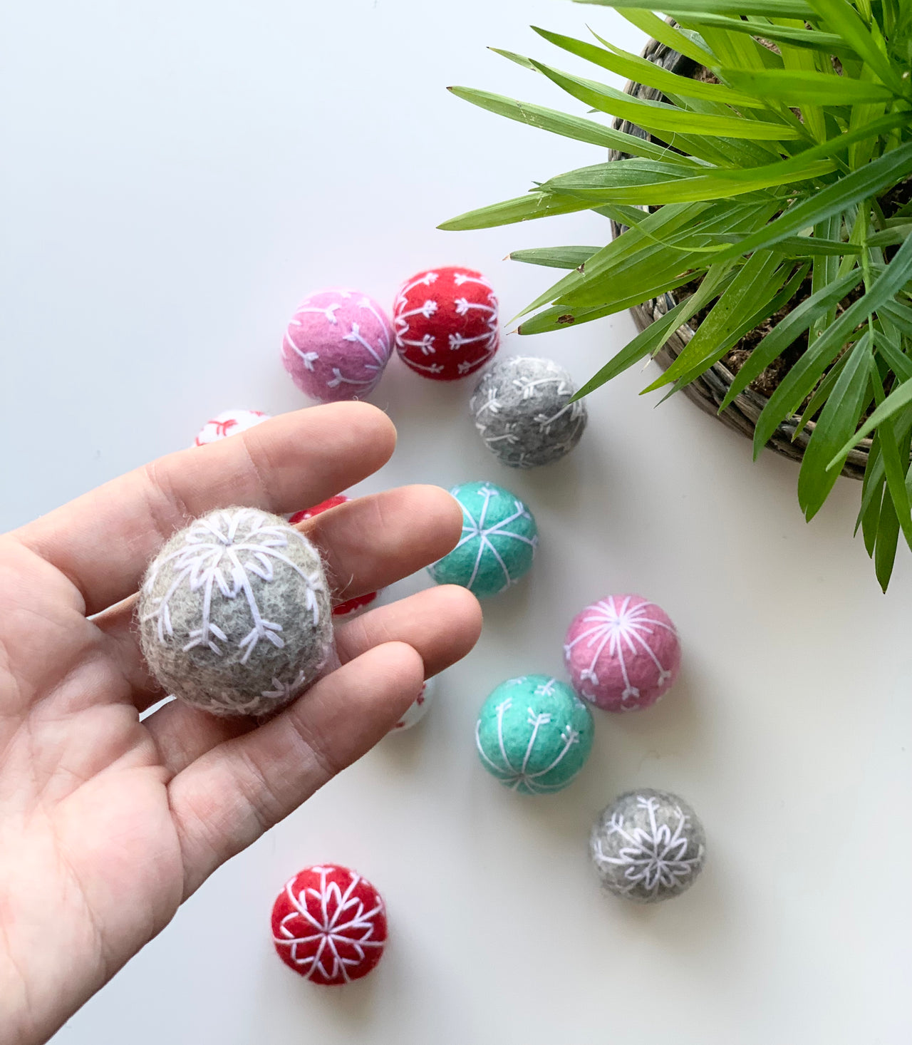 Hand holding a grey felt snowflake ball with assorted red, pink, teal, and grey felt balls and green plant on white surface