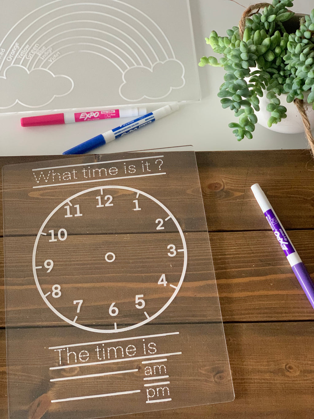 Clear acrylic dry erase tracing board with clock face and time practice lines on wooden table with markers and plant nearby