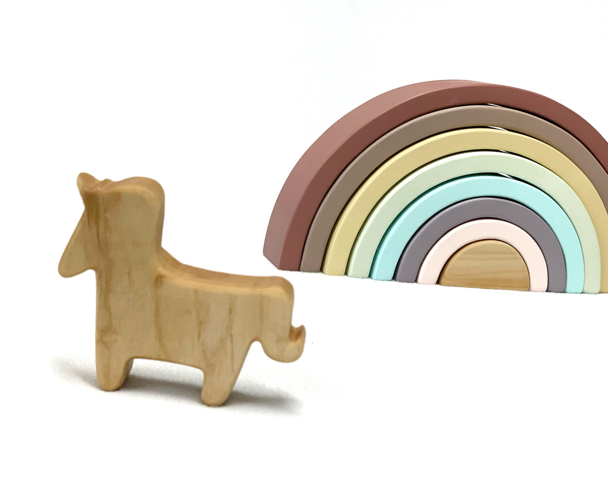 Small natural wooden unicorn toy figurine standing next to pastel-colored wooden rainbow stacking toy on white background