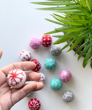 Hand holding single white felt snowflake ball with red embroidery and other colorful snowflake balls on white surface near green plant