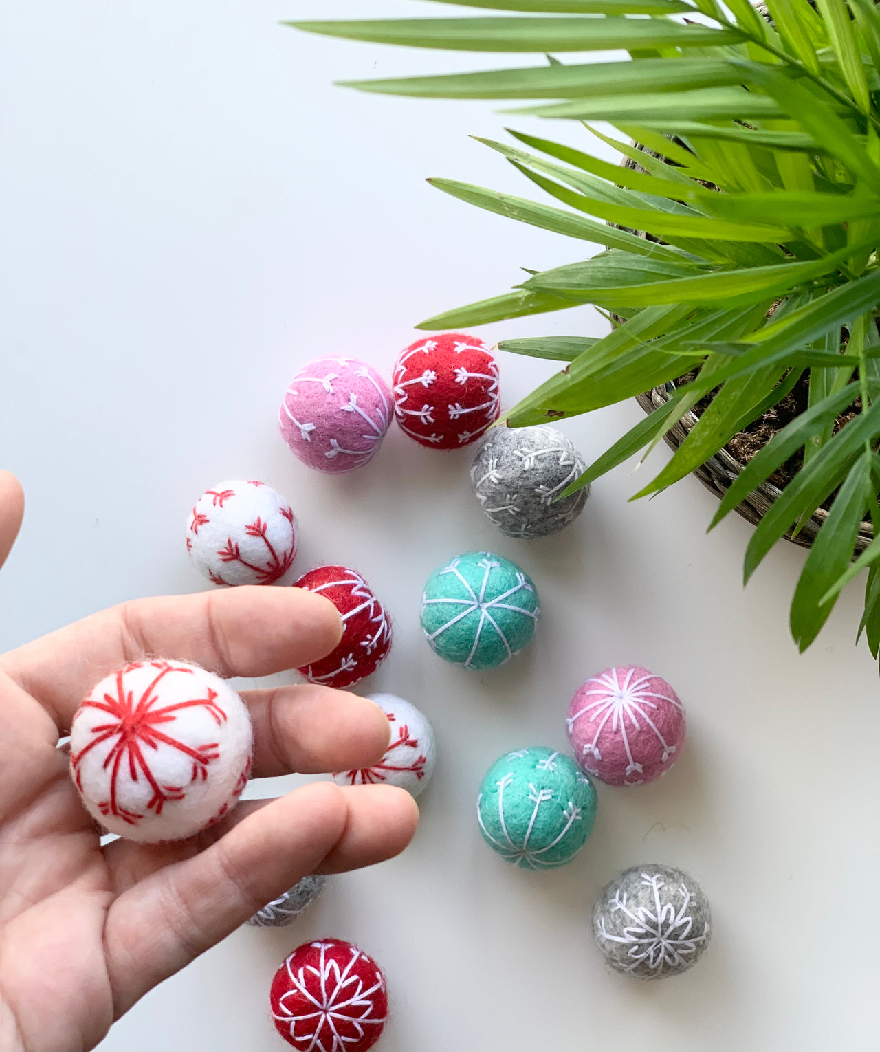 Hand holding single white felt snowflake ball with red embroidery and other colorful snowflake balls on white surface near green plant