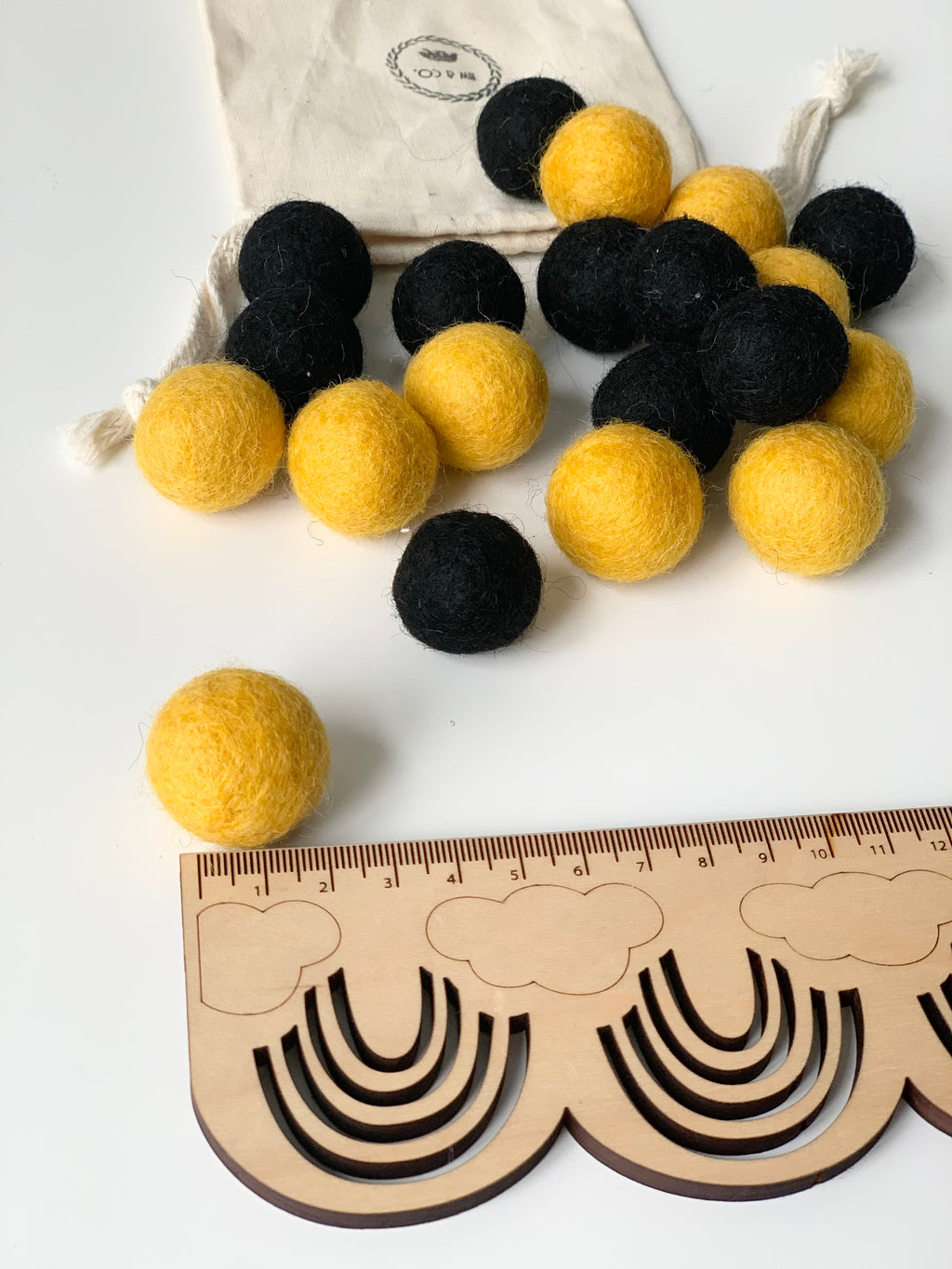 Yellow and black felt balls scattered on white surface near wooden rainbow ruler and white drawstring bag