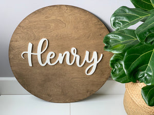 3D round wooden nursery sign with white cursive name lettering on natural wood grain background next to green plant